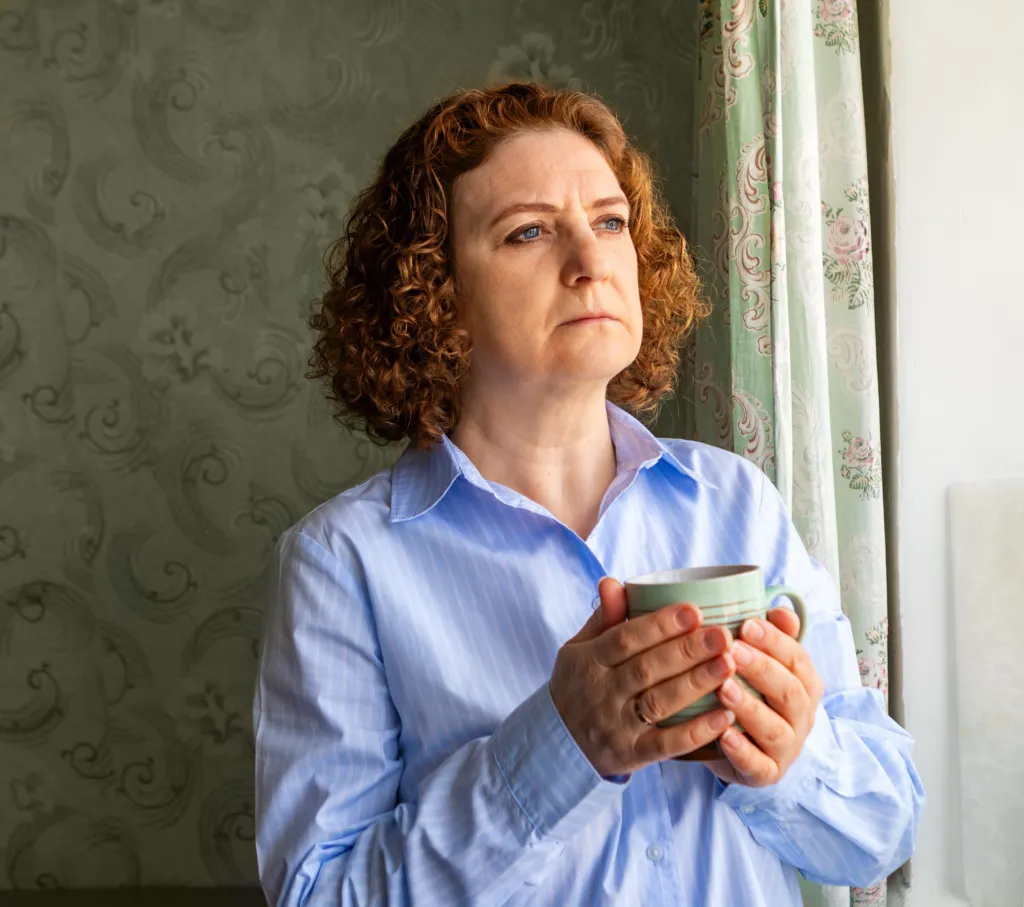 A woman holding a mug of tea, looking thoughtfully out of a window